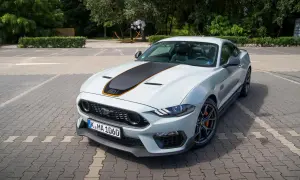 Biały Ford Mustang Mach 1 z czarnym pasem na masce i pomarańczowymi akcentami, zaparkowany na kostce brukowej.