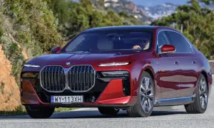 Czerwone BMW 740d z eleganckim czarnym dachem sunie po krętej drodze wśród zielonych wzgórz.