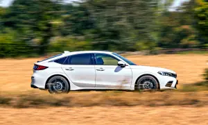 Biały Honda Civic 2025 pędzi przez suche pole, zostawiając za sobą smugi kurzu i rozmazane drzewa.