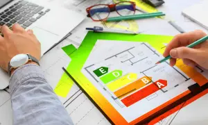 Rysunek domu z klasyfikacją energetyczną EPC. Ręka z ołówkiem analizuje plan, obok laptop i okulary.