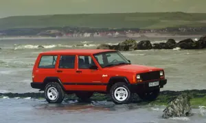 Czerwony Jeep ZJ na kamienistej plaży, z widokiem na morze i wzgórza w tle.
