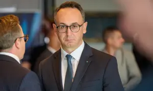 Minister infrastruktury w okularach i garniturze, z zieloną koniczynką na klapie marynarki, rozmawia z kimś na tle innych osób.