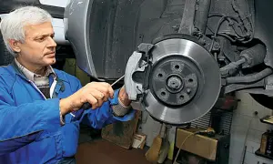 Mechanik w niebieskim kombinezonie naprawia hamulce, wymieniając klocki. W tle widać elementy układu kierowniczego i pompowtryski.
