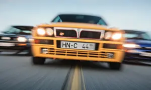 Żółta Lancia Delta Integrale w centrum, ścigana przez niebieski i czarny samochód.