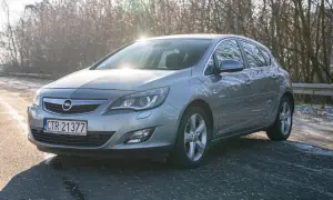 Szary Opel Astra 2011 na drodze, z drzewami w tle i słońcem odbijającym się od karoserii.