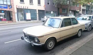 Klasyczny beżowy BMW E10 z lat 70. stoi zaparkowany przy ulicy, obok nowszego srebrnego samochodu.