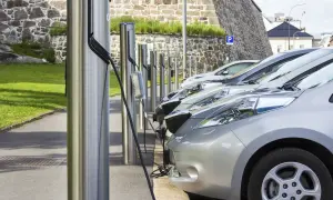 Rząd samochodów elektrycznych podłączonych do ładowarek na tle kamiennego muru.