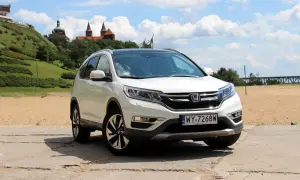 Biała Honda CR-V 2.0 benzyna na tle piaszczystego brzegu i zielonego wzgórza z budynkami.