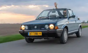 Mężczyzna w okularach przeciwsłonecznych macha ręką z kabrioletu Fiat Ritmo. Jazda po drodze o zachodzie słońca.