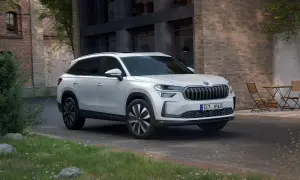 Biały Skoda Kodiaq 2025 zaparkowany przed ceglanym budynkiem, z zielenią i stolikiem z krzesłami w tle.