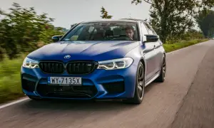 Niebieskie BMW M5 F90 pędzi po wiejskiej drodze, kierowca skupiony na trasie.