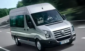 Srebrny Volkswagen Crafter jedzie po krętej drodze wśród drzew. Kierowca w białej koszuli prowadzi auto.