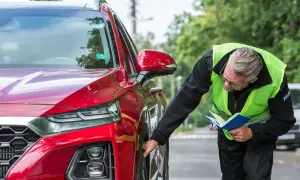 Policjant w kamizelce odblaskowej sprawdza oponę czerwonego auta, notując coś w niebieskiej książeczce. To część jego pracy, być może związanej z art. 97 kw.
