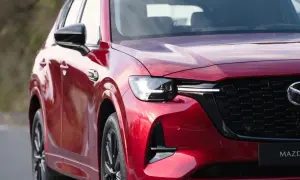 Błyszczący, czerwony lakier nowej Mazdy CX-60 odbija niebo. Widoczny fragment maski, reflektor i charakterystyczny grill.