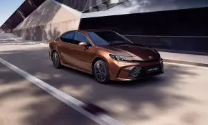 Brązowa Toyota Camry Hybrid 2024 pędzi po drodze, otoczona nowoczesną architekturą i cieniami drzew.