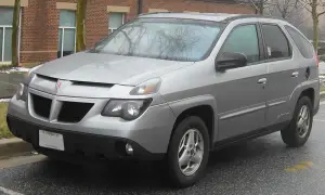 Srebrny Pontiac Aztek zaparkowany na mokrym asfalcie, z ceglanym budynkiem w tle.