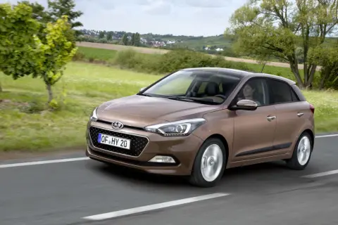 Brązowy Hyundai i20 jedzie po drodze, za nim zielone pola i domy. Sprawdź atrakcyjną hyundai i20 cena!