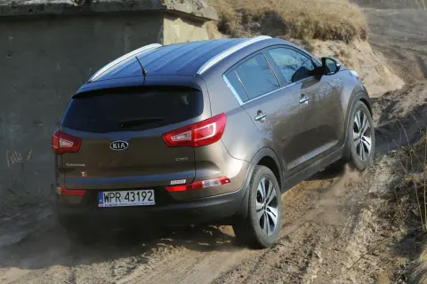 Brązowy Kia Sportage 2014 zjeżdża po piaszczystej ścieżce, unosząc kurz.