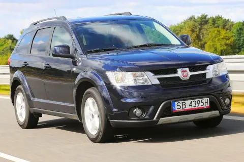Ciemnoniebieski Fiat Freemont jedzie po drodze, otoczony zielenią i błękitnym niebem.