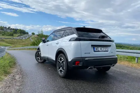 Biały Peugeot 2008 GT na mokrej drodze, z widokiem na krętą trasę i zielone wzgórza.