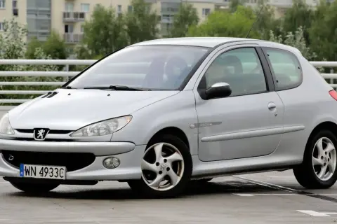 Srebrny Peugeot 206 Plus z tablicą rejestracyjną WN 4933C stoi na mokrym parkingu.