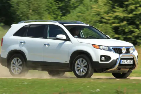 Białe Kia Sorento 1 zderza się z kurzem na drodze gruntowej, jadąc przez zielone pola.