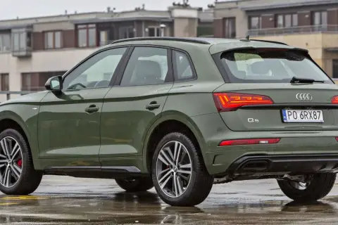 Zielony Audi Q5 Sportback na mokrym parkingu, z budynkami w tle.