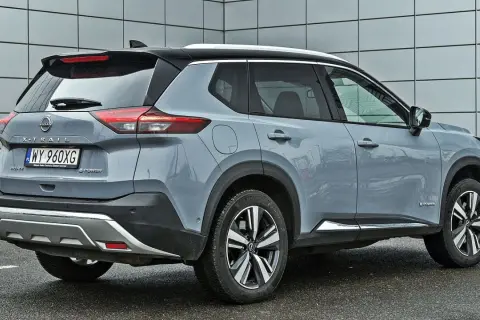 Szary Nissan X-Trail e-POWER z czarnym dachem i srebrnymi akcentami stoi na mokrym parkingu przed nowoczesnym budynkiem.