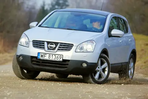 Srebrny Nissan Qashqai 1. pokonuje zakręt na piaszczystej drodze, wzbijając kurz.