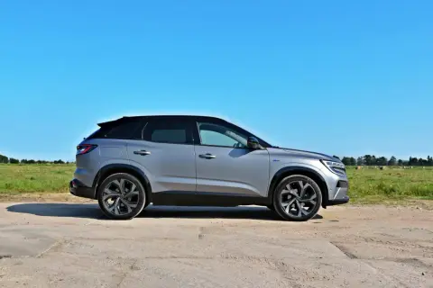 Srebrny Renault Austral z czarnym dachem stoi na polnej drodze, w tle zielone pole i błękitne niebo.
