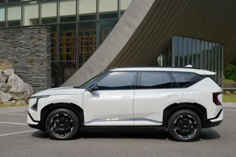Nowy, biały suv Kia EV9 stoi zaparkowany przed nowoczesnym budynkiem z kamienną fasadą i dużymi oknami.