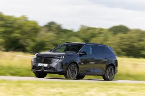 Ciemnoszary Peugeot 5008 jedzie po drodze wśród zielonych pól i drzew.