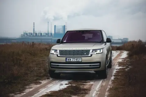 Złoty Range Rover Autobiography na błotnistej drodze, w tle przemysłowy krajobraz z kominami fabrycznymi.