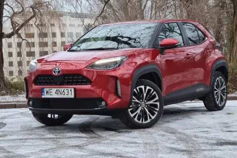 Czerwony Toyota Cross Hybrid na tle drzew i budynku. Auto stoi na ośnieżonej nawierzchni.