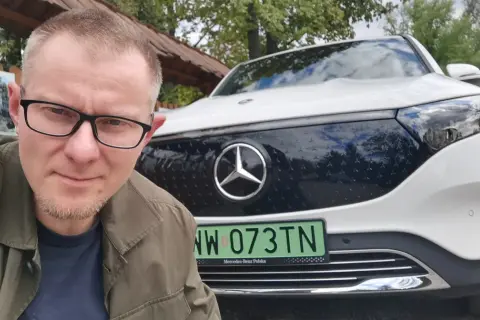 Mężczyzna w okularach pozuje do selfie na tle białego Mercedesa EQB.