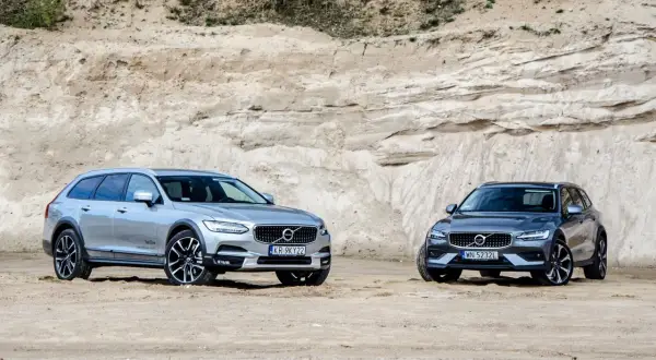 Dwa samochody Volvo Cross Country, srebrny i grafitowy, stoją na piaszczystym podłożu przed piaskową ścianą.