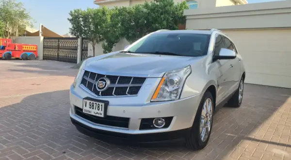 Srebrny Cadillac SRX z Dubai stoi na podjeździe przed domem. Obok widać bramę i fragment czerwonej zabawki.