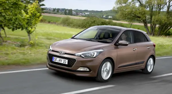 Brązowy Hyundai i20 jedzie po drodze, za nim zielone pola i domy. Sprawdź atrakcyjną hyundai i20 cena!
