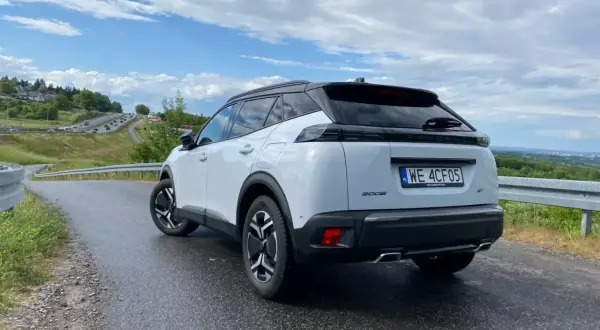 Biały Peugeot 2008 GT na mokrej drodze, z widokiem na krętą trasę i zielone wzgórza.