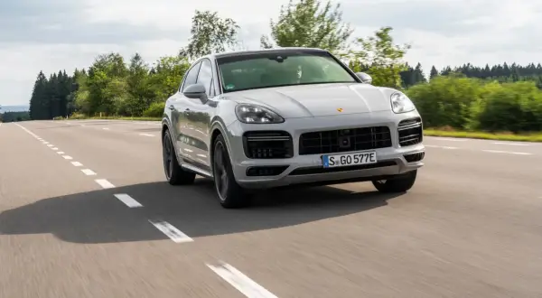 Srebrny Porsche Cayenne 2025 sunie po asfaltowej drodze, otoczony zielenią lasów.