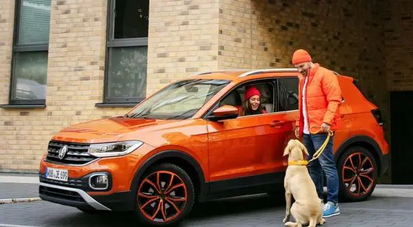 Pomarańczowy mini suv z kobietą w środku i mężczyzną z psem na smyczy.