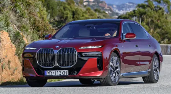 Czerwone BMW 740d z eleganckim czarnym dachem sunie po krętej drodze wśród zielonych wzgórz.
