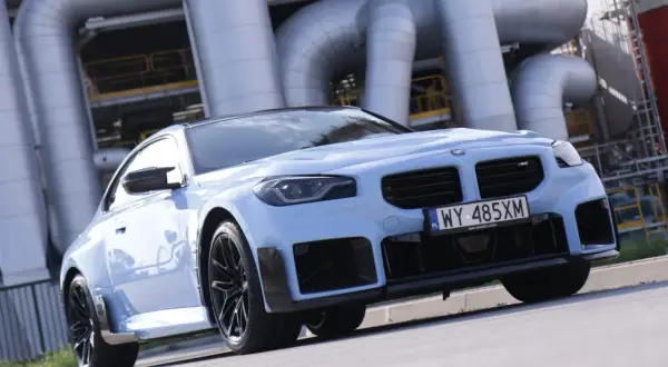Jasnoniebieskie BMW M2 z czarnymi felgami i spoilerami, na tle przemysłowych rur. Sprawdź aktualną bmw m2 cena.