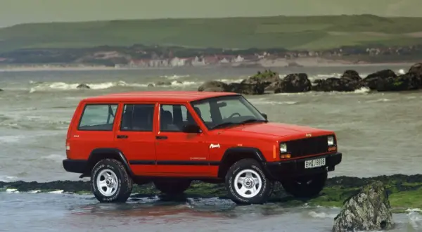 Czerwony Jeep ZJ na kamienistej plaży, z widokiem na morze i wzgórza w tle.