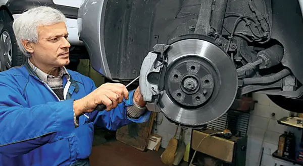Mechanik w niebieskim kombinezonie naprawia hamulce, wymieniając klocki. W tle widać elementy układu kierowniczego i pompowtryski.