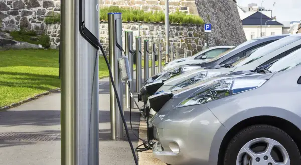 Rząd samochodów elektrycznych podłączonych do ładowarek na tle kamiennego muru.