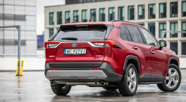 Czerwony Toyota RAV4 2020 Hybrid na mokrym parkingu, z nowoczesnym budynkiem w tle.
