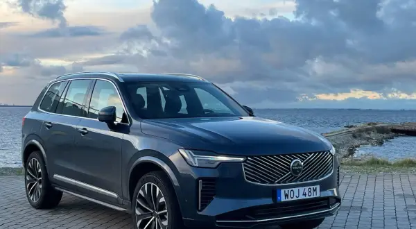 Elegancki, ciemnogranatowy Volvo XC90 2025 zaparkowany nad morzem, z chmurami na niebie.