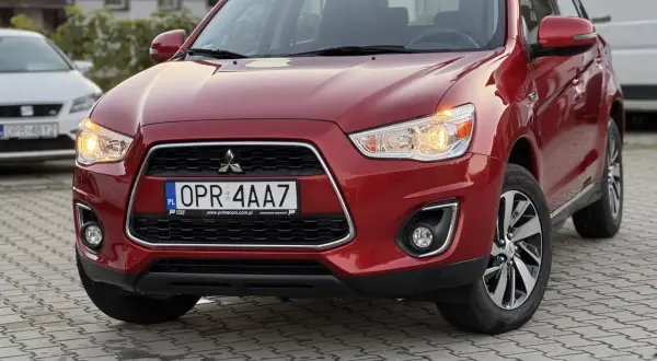 Czerwony Mitsubishi ASX 1.6 benzyna z tablicą rejestracyjną OPR 4AA7 zaparkowany na kostce brukowej.