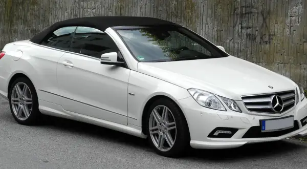 Biały kabriolet Mercedes W207 z czarnym dachem stoi na ulicy obok betonowej ściany.